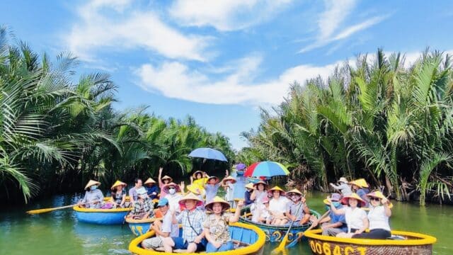 Tourists in round basket boats paddle through Hoi An’s green river, lined with palm trees under a bright blue sky.