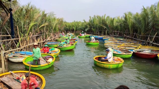 People ride colorful round basket boats in Hoi An on a narrow river, surrounded by lush green palm trees under a partly cloudy sky.