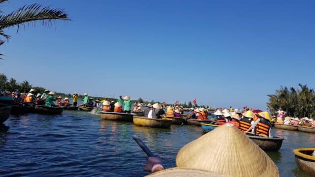 People wearing conical hats paddle round basket boats on a calm river in Hoi An, surrounded by lush greenery under a clear blue sky.