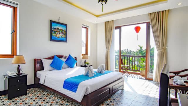 Bright Hoi An hotel room with blue accents, balcony, large windows, and two towel elephants on the bed.
