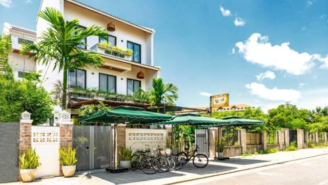 Modern white three-story building with balconies and green awnings in Hoi An, palm trees, and bicycles parked out front on a sunny day.