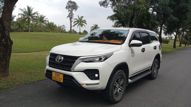A white Toyota SUV with a yellow license plate parked on a paved road in Hoi An, next to grass and trees.