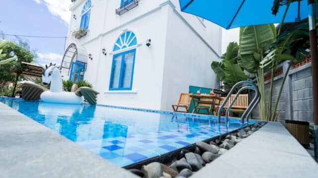 A white house with blue trim in Hoi An, a pool with a unicorn float, patio set, and tropical plants under a blue umbrella.