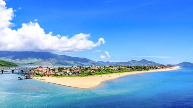 Coastal town of Hoi An with sandy beach, blue water, a bridge, and green mountains under a clear blue sky.