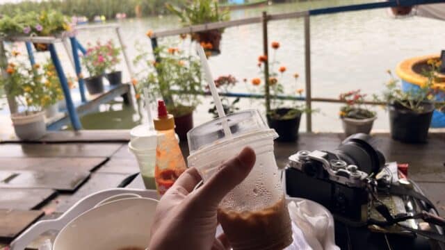 A hand holds an iced drink by a lakeside table in Hoi An, surrounded by food, flowers, a camera, water, and lush trees in the background.