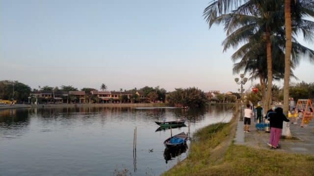 People stand by a riverside path in Hoi An, with boats in the water and palm trees lining the edge at sunset.