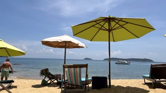 Beach chairs and umbrellas on Hoi An’s sandy shore, with a man standing and a boat on the ocean under a sunny blue sky.