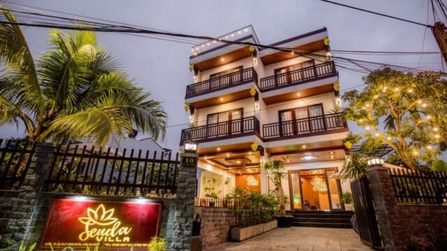 Three-story villa in Hoi An with balconies, warm lights, palm trees, and a glowing Senda Villa sign at the entrance.