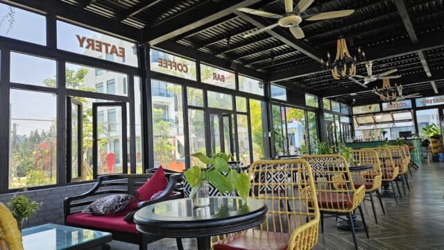 A bright, modern Hoi An cafe with wicker chairs, glass tables, lush plants, and large windows letting in natural light.
