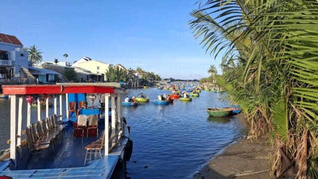 Colorful round boats and a larger boat float on Hoi An’s calm river, beside houses and palm trees under a clear blue sky.