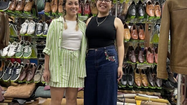 Two women smiling in a Hoi An leather goods store, surrounded by shoes, bags, and jackets on shelves and racks.