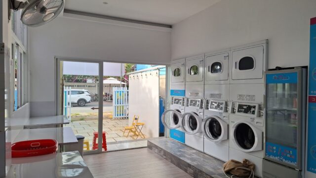 Modern laundromat in Hoi An with washers, dryers, folding tables, a fan, and a view outside through a glass door.
