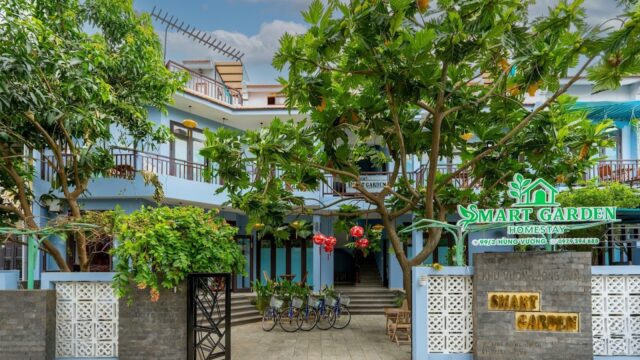 The entrance of Smart Garden Homestay in Hoi An features a blue building, leafy trees, bicycles, and a bright green sign above the gate.