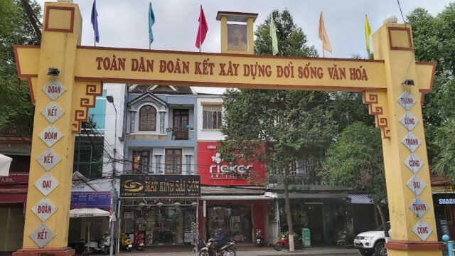 Yellow archway with Vietnamese text, Hoi An flags on top, and shops and trees visible in the background.