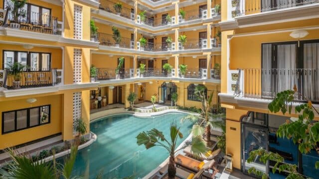 Open-air Hoi An hotel atrium with a central swimming pool, yellow walls, balconies, and potted plants on each floor.