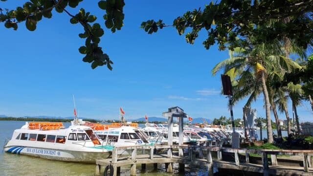 Several white passenger boats docked at a wooden pier in Hoi An, with palm trees and blue sky in the background.