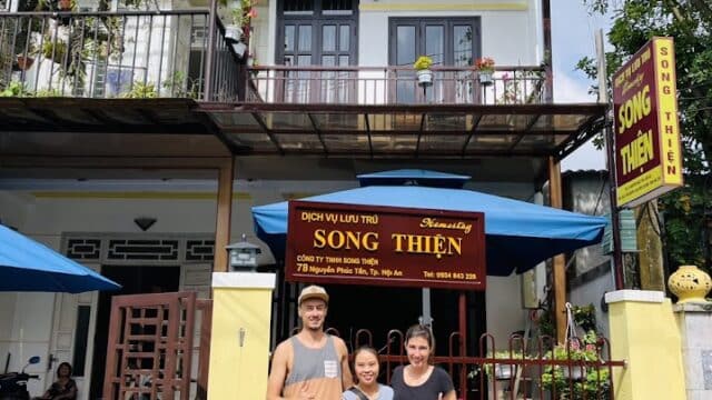 Three people smiling and posing in front of a Hoi An guesthouse with Song Thien signs and plants by the entrance.