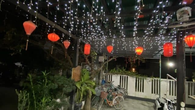 Red lanterns and string lights glow above bicycles and green plants in a Hoi An outdoor area at night.