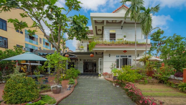A two-story white house in Hoi An with a garden, potted plants, and a tiled walkway under a blue sky.