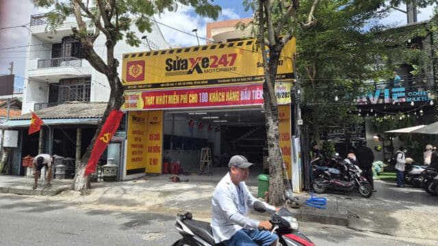 A man rides a motorbike past a yellow Sửa Xe 247 repair shop on a sunny Hoi An street.