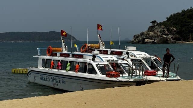 Two white boats with life vests and flags docked on a sandy Hoi An beach beside clear blue water and distant hills.