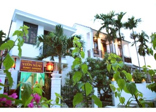 White two-story house with palm trees and green plants in Hoi An, sign reads Vuon Nang Sunny Garden at entrance.