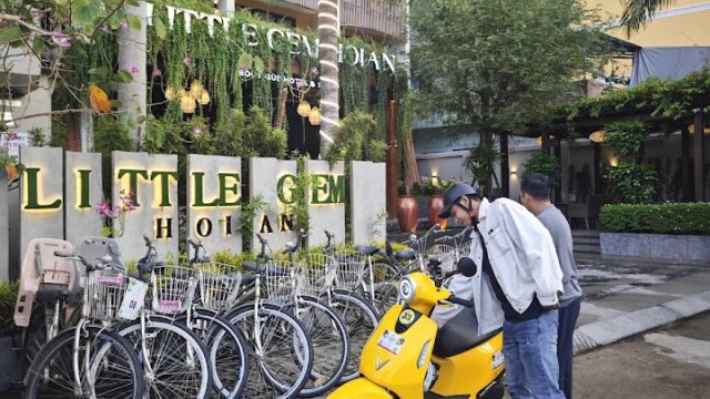 Two people stand by a yellow scooter in front of the Little Gem Hoi An hotel sign, with bicycles lined up nearby in Hoi An.