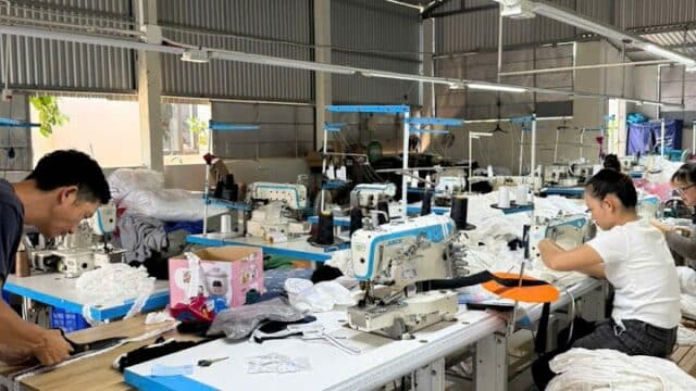 People working at sewing machines in a Hoi An garment factory, surrounded by fabric, clothing pieces, and equipment on the tables.