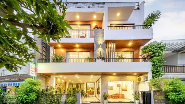 Modern three-story Hoi An house with large balconies, warm lighting, plants, floor-to-ceiling windows, and lush greenery all around.