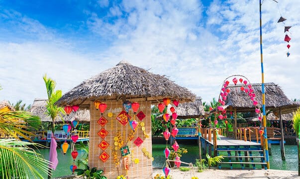 Tropical huts with thatched roofs and colorful lanterns line a clear blue waterway in Hoi An under a bright, partly cloudy sky.