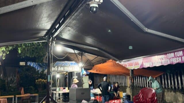 Outdoor street food stall at night in Hoi An, under large tents with red chairs, tables, and people preparing delicious food.