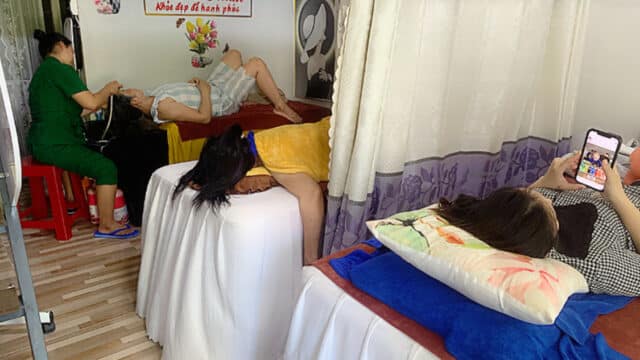 Three women enjoy spa treatments in a cozy, curtained Hoi An room; one uses her phone while another gets her hair washed.
