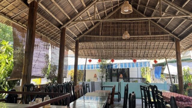 Open-air Hoi An restaurant with wooden roof, empty tables, bamboo shades, and natural light streaming through lush plants outside.