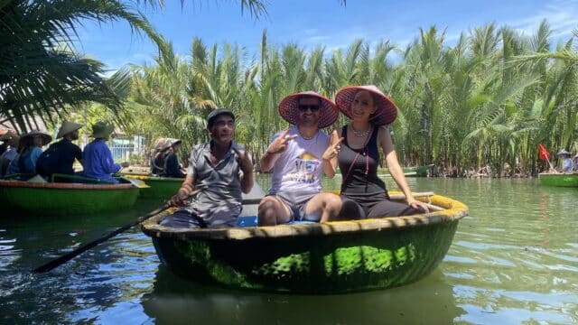 Three people smile in a round basket boat on Hoi An’s river, surrounded by lush palms under a bright blue sky.
