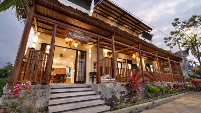 A cozy wooden guesthouse in Hoi An with a wide porch, lush plants, and warm lighting glowing at dusk.