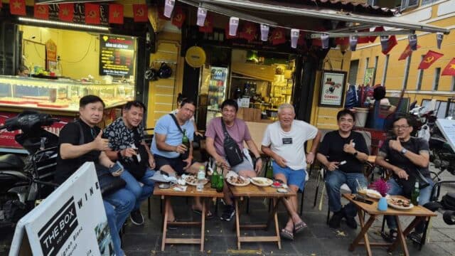 Seven men sit at small tables outside a yellow café in Hoi An, smiling and giving thumbs up with food and drinks in front of them.