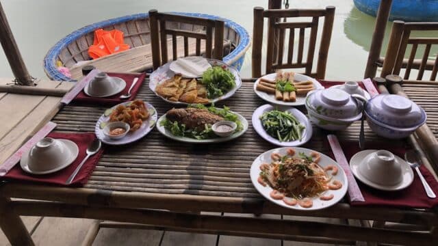 A bamboo table by the water in Hoi An, set with various Vietnamese dishes, bowls, plates, and fresh greens.