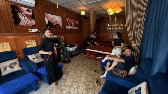 Four people enjoy a cozy Hoi An massage spa with blue chairs, wooden walls, and calming decor—one relaxes as others chat nearby.