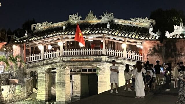 The historic Japanese bridge in Hoi An, Vietnam, glows at night with a Vietnamese flag and visitors nearby.