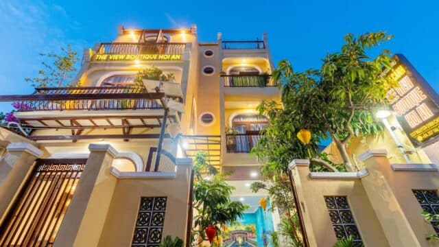 A brightly lit Hoi An boutique hotel with balconies and greenery, viewed from the gated entrance at dusk.