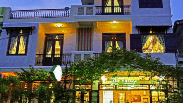 A warmly lit, two-story building with large windows and a sign reading The Village at the entrance, inspired by Hoi An charm.