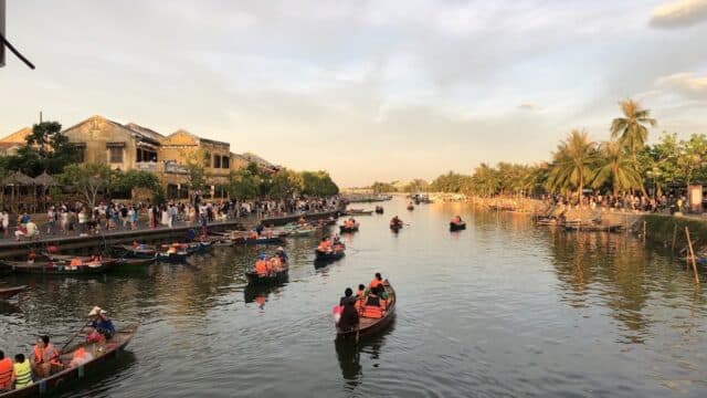 People in boats row along a river lined with palm trees, crowds, and historic Hoi An buildings at sunset.