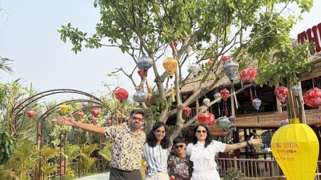 Four people stand smiling under a tree with colorful lanterns, enjoying a bright Hoi An day near a wooden building and yellow lantern.