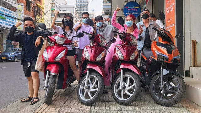 Six people wearing masks in Hoi An stand behind three red and one orange scooter, raising their hands on a city sidewalk.