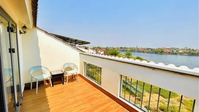 Sunny Hoi An balcony with two chairs, small table, wooden floor, and river view framed by houses and lush greenery.