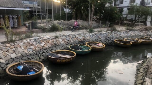Round basket boats are lined up along a canal in Hoi An, with houses and palm trees in the sunset background.