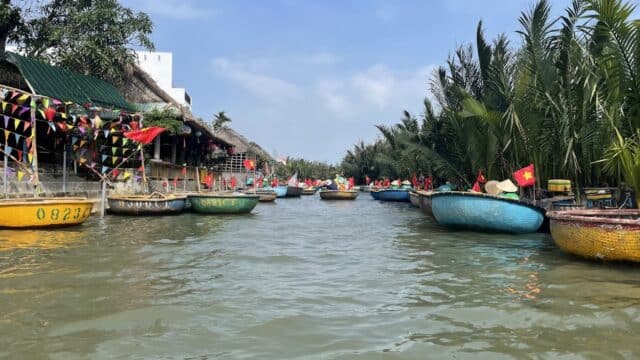 Round basket boats with flags float on a river in Hoi An, lined with palm trees and buildings under a clear blue sky.