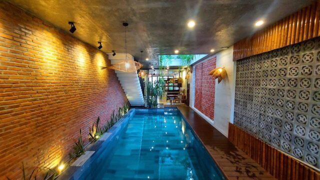 Indoor pool with brick walls, modern lighting, and lush plants evokes a Hoi An vibe, leading to a staircase and seating area.