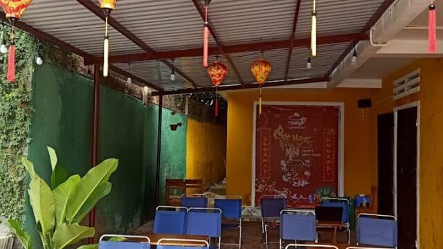 Outdoor seating area with blue chairs, wooden tables, and red lanterns hanging from a covered roof, evoking Hoi An charm.