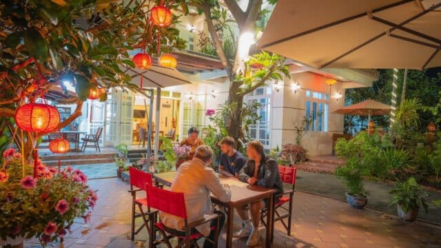 Four people sit at a table in a brightly lit garden patio, surrounded by plants and Hoi An-inspired red lanterns at night.